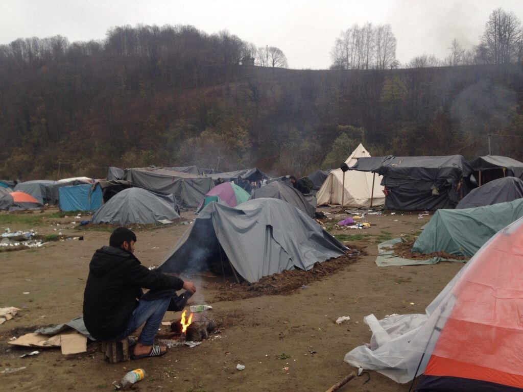 Život v utečeneckom tábore, muž sedí pri ohni a okolo neho je množstvo stanov, v ktorých prespávajú ľudia. 