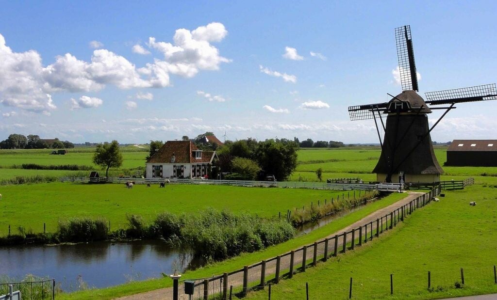 Holandská krajina so zelenou trávou, jazierkom, domom, modrou oblohou a veterným mlynom 