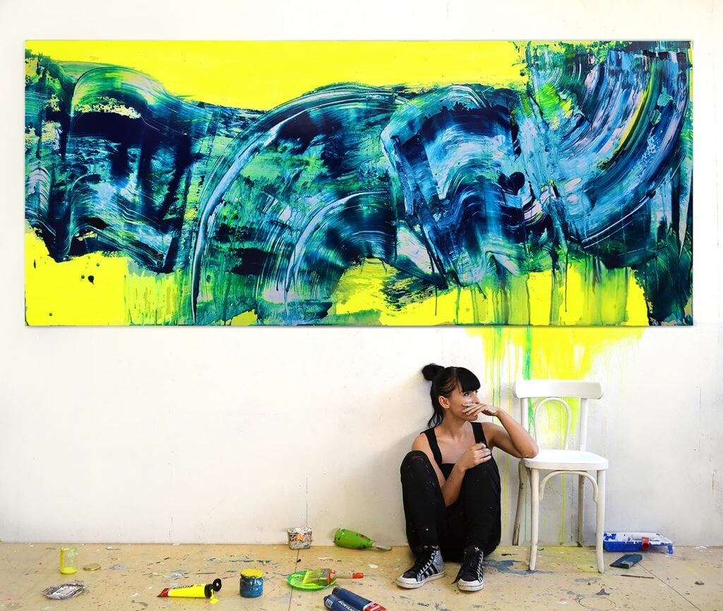 Maliarka Hula pred svojím plátnom, na ktorom je abstraktná maľba, ktorá sa nesie v znamení modrej a žltej. 